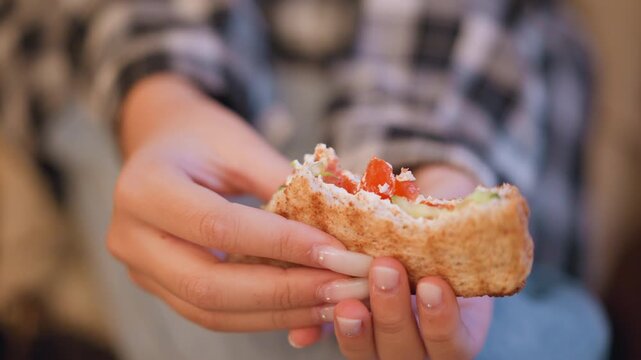 Close up of woman holding and eating half eaten pita bread with tomato filling. subject hands wearing checkered clothing gently cradle food item showcasing detailed nail texture and soft