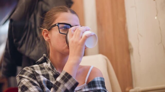 Close up of woman drinking from white cup while wearing glasses and casual clothing. scene shows soft indoor lighting with warm tones featuring wooden elements in background and minimal