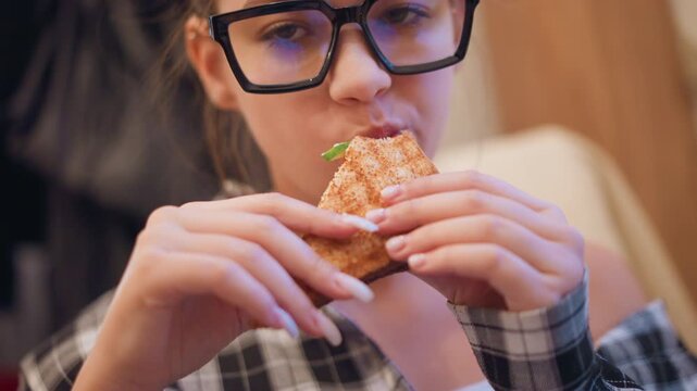 Close up of young woman with long hair and glasses eating food. Woman wears black framed eyeglasses has manicured nails painted white on natural color and is dressed in plaid clothing