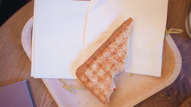 Close up of partially eaten toasted sandwich on white napkins placed near open notebook and wooden table. scene features soft lighting with warm tones highlighting texture of bread crumbs