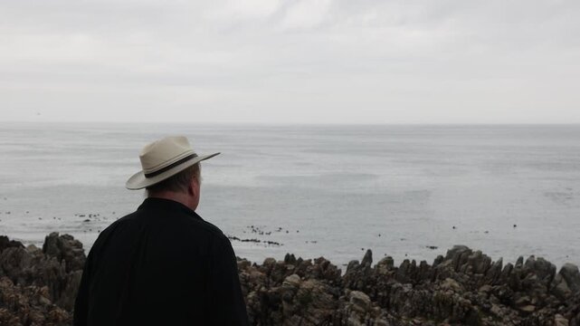 man wearing hat overlooking stormy dawn clouds over rocky coastline of DeKelders, South Africa