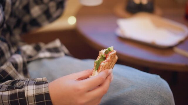 Close up of young woman holding and eating sandwich with visible lettuce tomato in indoor setting. scene includes soft lighting warm tones wooden table background elements like cup plate