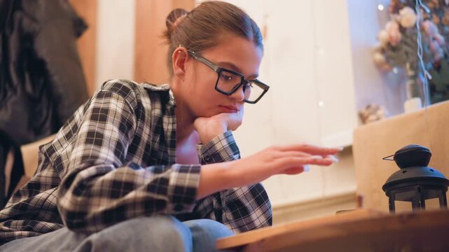 Close up of young woman with glasses resting her chin on one hand while seated indoors wearing casual attire. Woman appears contemplative or focused in soft lighting surrounded by neutral