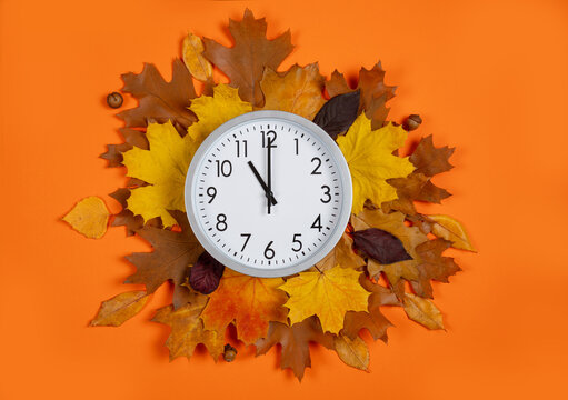 Top view: white round analog clock sits in the central of autumn leaves against a bright orange background. Conceptual flat lay composition for transition of the fall season, time management.