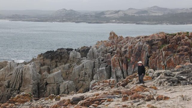 man wearing hat looking at stormy dawn clouds over rocky coastline of DeKelders, South Africa