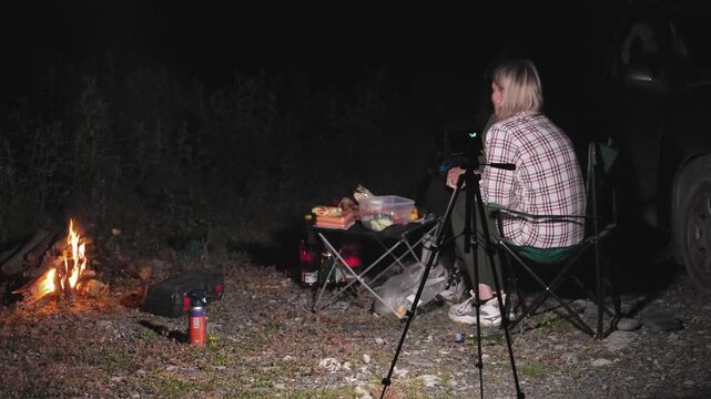 Blonde woman wearing a plaid shirt sitting in a camping chair, filming a crackling bonfire with a camera on a tripod during a night car camping trip while preparing food on a small table