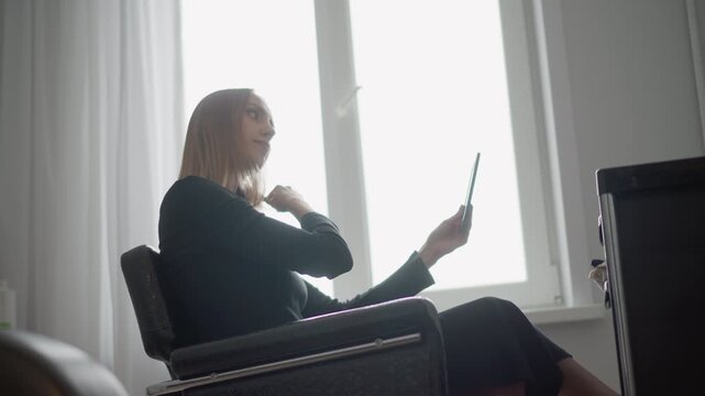 Woman in black outfit sits on chair using tablet and brushing hair near window with white curtains. Close up shows personal grooming activity indoors with soft natural light from large