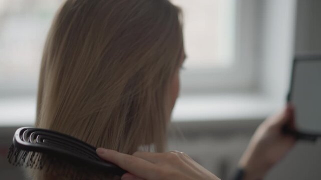 Close up of woman brushing long brown hair with smooth strokes while holding small mirror in one hand. action is performed indoors likely near window providing soft natural light focusing