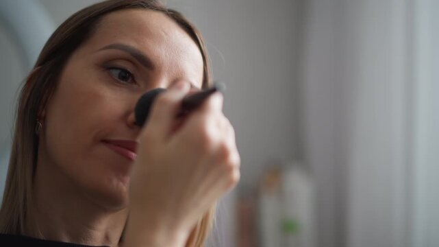 Close up of woman applying makeup with soft brush in bathroom setting. Woman holds and moves black powder brush near her face neck visible as woman focuses on grooming routine. Background