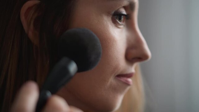 Close up of woman applying makeup with soft brush to face in calm indoor setting. Makeup application process shown focusing on hand and tool interaction against blurred background. Lighting