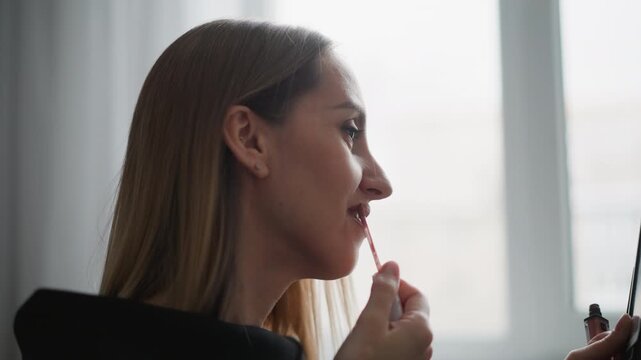 Close up of woman applying lip gloss while looking into handheld mirror. scene shows steady focus on application process with soft natural light filling indoor setting emphasizing personal