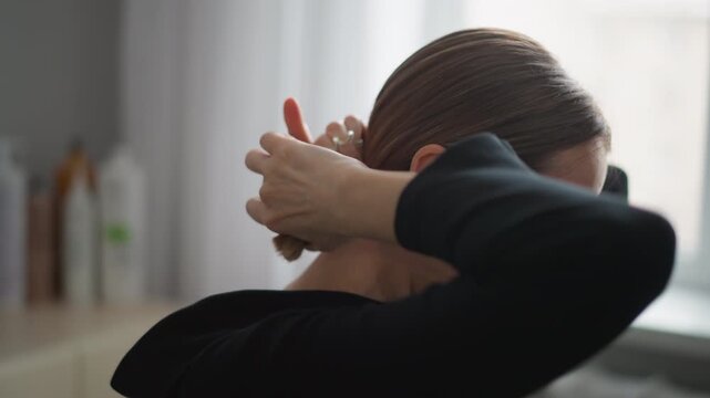 Close up of woman styling her hair with hands gathering and securing it into elegant low ponytail. Hair is smooth brown in texture while wearing black clothing set against soft indoor