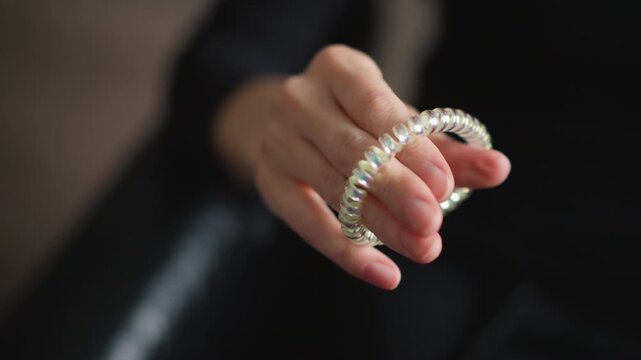 Close up of hand holding and manipulating iridescent coiled hair tie against dark background. Hand moves slightly to display flexible accessory emphasizing its shiny texture and form. Soft