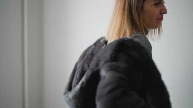 Close up of woman putting on and adjusting black fur coat over gray turtleneck sweater. subject moves slightly revealing texture details of both clothing materials against plain light