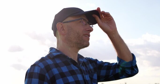 Man in cap and glasses adjusts visor outdoors. Man looks across open landscape under cloudy sky. Smiling man enjoys peaceful outdoor moment. Outdoor lifestyle footage suits nature campaigns.