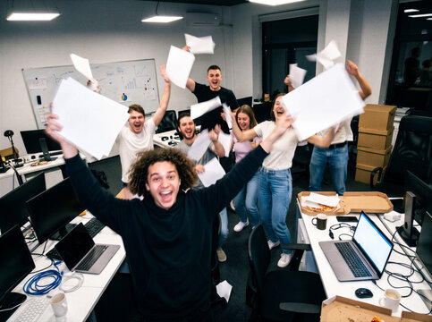Ecstatic startup team throwing paper in the air to celebrate project completion in a messy modern office