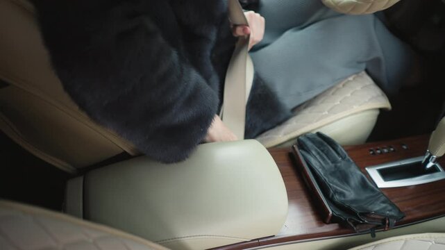 Close up of woman in dark fur coat fastening her seatbelt inside automobile. interior features beige leather seats wooden center console with black gloves and gear shift visible soft