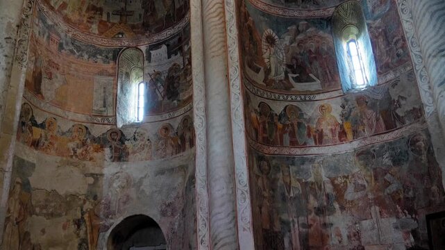 The interior of ancient stone Orthodox church of Nikortsminda in Georgia with weathered Byzantine frescoes of saints and biblical scenes on domes and curved walls, sunlight and candlelight flood nave