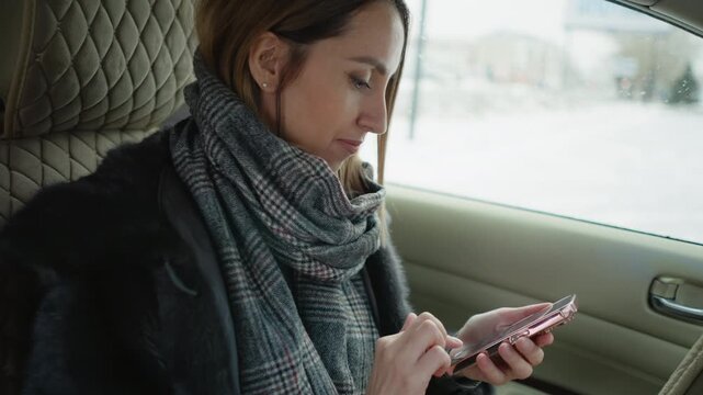 Close up of woman using smartphone in car seat wearing plaid scarf and dark coat. Scene shows her interacting with phone transitioning from typing to holding it near face while seated
