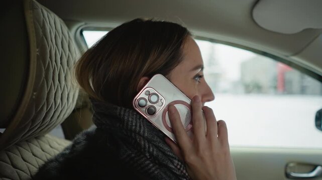 Close up of woman talking on smartphone while sitting in car seat. Hand holding phone to ear wearing patterned scarf and dark coat. Car interior with quilted headrest visible window