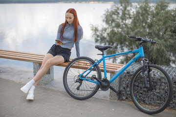 Fototapeta premium Young woman resting on a waterfront bench beside her bicycle while checking her smartphone. Cycling, leisure and outdoor lifestyle concept.