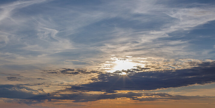 Sun shining through layered clouds at sunset, creating a dramatic sky with warm golden light, soft blue tones and a peaceful atmospheric landscape.