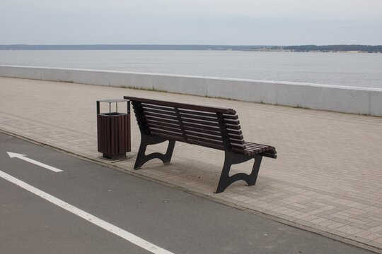 Empty wooden bench beside a waterfront promenade overlooking calm water. Minimal outdoor scene for relaxation, travel, recreation and peaceful lifestyle concepts.