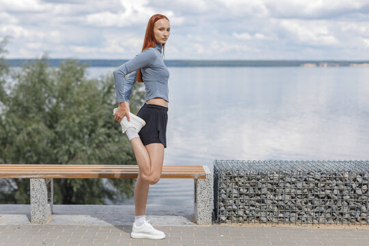 Young woman stretching her quadriceps beside a waterfront before a run. Outdoor warmup and preparation for exercise in a peaceful summer setting.
