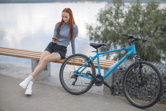 Young woman resting on a waterfront bench beside her bicycle while checking her smartphone. Cycling, leisure and outdoor lifestyle concept.