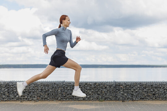 Young woman sprinting along a waterfront promenade under a cloudy summer sky. Outdoor jogging, endurance training and healthy active lifestyle concept.