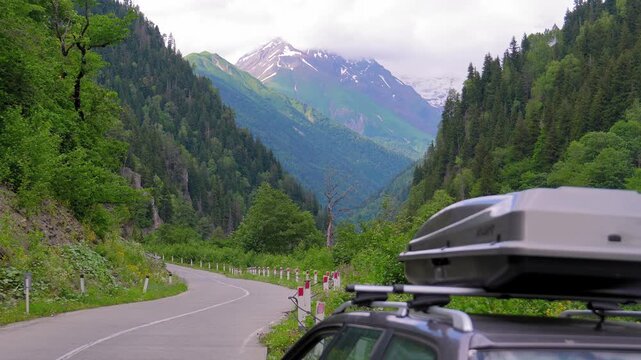 Adventurous road trip concept with a car and its rooftop cargo box parked on a scenic mountain pass overlooking a beautiful green valley and snowy peaks during a summer vacation journey georgia