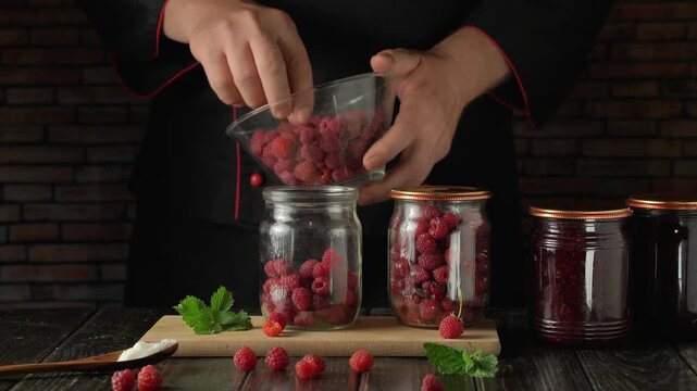 Chef in black uniform fills glass jars with fresh raspberries from a bowl, surrounded by mint leaves and wooden utensils on a rustic wooden table against a brick wall backdrop