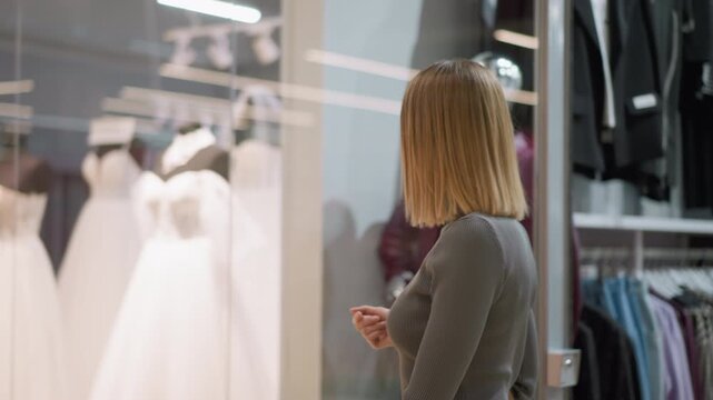 Woman with shoulder length hair browses clothing store observing mannequins and hanging garments. Woman wears fitted gray top while holding her hand near chest level in indoor retail