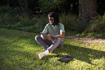 Adult African man sitting cross-legged on grassy lawn in park using tablet and portable solar panel © WavebreakMediaMicro