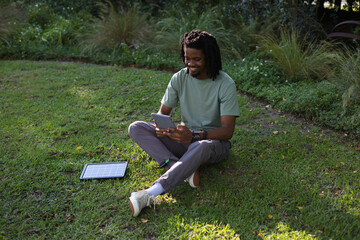 Obraz premium African man sitting cross-legged on grassy lawn, holding tablet, using portable solar panel nearby