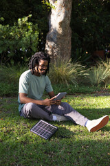 African American man sitting on lawn under tree, holding tablet charging via solar panel cable © WavebreakMediaMicro