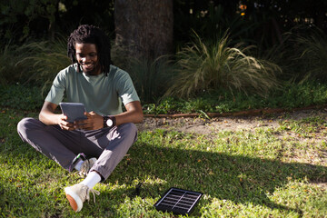 African American man wearing tee sitting on lawn holding tablet charging via solar panel with cable © WavebreakMediaMicro