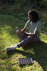 Man sitting on sunlit lawn holding tablet, charging with portable solar panel and basketball nearby © WavebreakMediaMicro