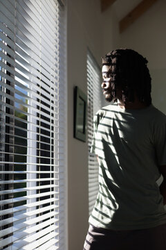 African American man standing in room by window with blinds, sunlight casting stripes, copy space