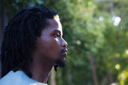 African man posing at left in park wearing light crew tee, dreadlocks and bokeh, copy space