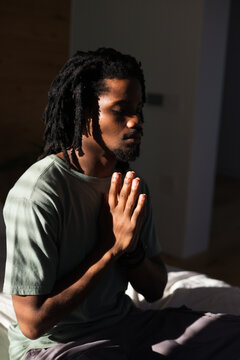 African man meditating on bed in quiet bedroom, wearing muted green Tshirt and beaded bracelet