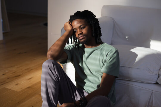 Adult African American man sitting on floor against sofa, showing watch, sunlight casting shadows