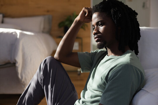 African American man 20s wearing green tee sitting on bedroom floor leaning against bed, copy space