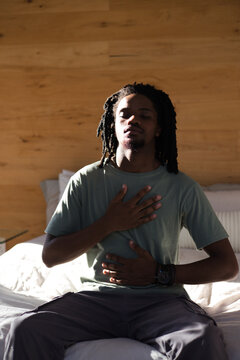 African American man sitting on bed in bedroom by wood paneling, wearing green tee and wristwatch