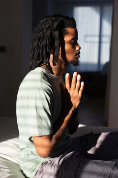 African American man sitting in bedroom pressing hands in green tee blinds casting shadow bands