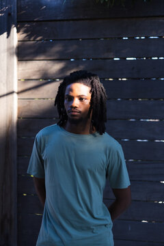Adult male standing centered in fenced yard wearing teal T-shirt, sunlight casting slat shadows
