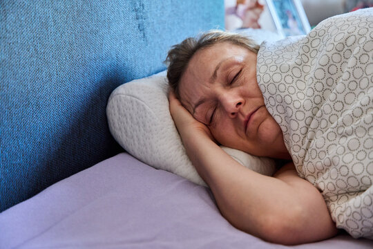 Middle-aged woman sleeping peacefully in her bed on an orthopedic foam pillow under a patterned blanket.