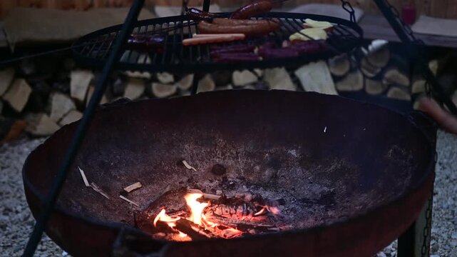 Video footage of sausages grilling over a campfire bowl on a tripod, ending with a person adding fresh firewood to the burning embers.