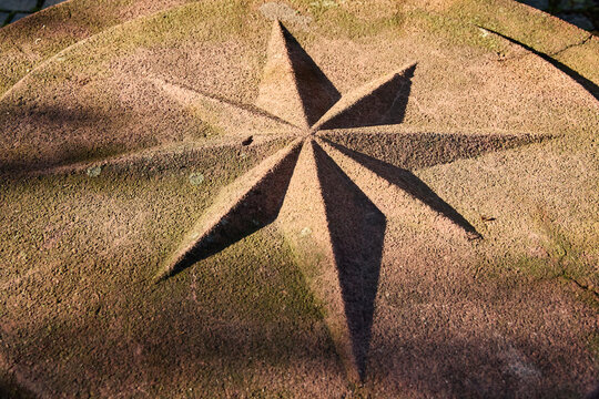 Close-up of a carved eight-pointed star or compass rose symbol on a round rough stone surface with sunlight casting shadows.