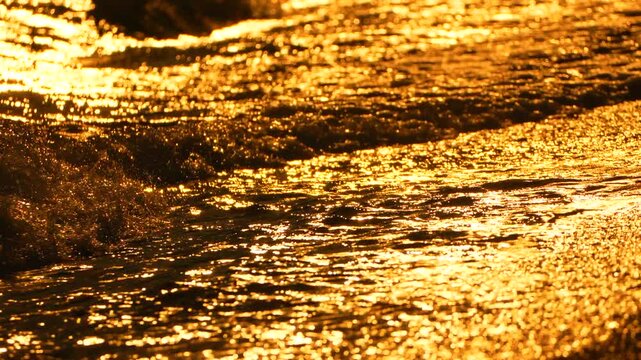 Ocean waves crash against the sandy shoreline at dusk, illuminating dark waters with intense yellow sunlit reflections. Static close-up shot.
